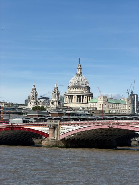 A view of St. Paul's Cathedral in London seen from across the River Thames, with a red and white bridge in the foreground. The clear blue sky highlights the historic dome and detailed façade of the c